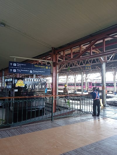 The image depicts an outdoor train station platform in Cangkring, Indonesia, under a pale green corrugated metal roof supported by reddish-brown metal beams and trusses. In the foreground, a dark green metal railing with decorative scrollwork separates the viewer's area from the train tracks. The floor is tiled with alternating light brown and greyish-brown rectangular patterns.
An overhead black sign with white arrows and text displays "Keamanan (Security)," "Pos Kesehatan (First Aid)," "Keberangkatan (Departure)," and "Ruang Menyusui (Nursery Room)." To the left, a black kiosk branded "ROTI MARYAM SALMAN" is positioned behind the railing.
Approximately five individuals are visible. In the mid-ground, to the right, a man in a blue polo shirt, dark trousers, and a yellow reflective safety vest leans on the railing, facing towards the tracks. Further back on the platform, another man in a beige uniform and yellow hard hat stands near the tracks. A long train with purple and silver livery is parked on multiple sets of tracks behind the railing. Some sections of the platform and tracks appear wet. Other partially obscured individuals are present further down the platform near the train.