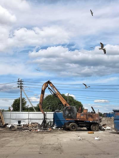This media file captures an outdoor scene, likely in a junkyard or industrial area, under a partly cloudy sky. The primary object of interest is a large, orange excavator with a claw attachment, positioned in the center of the frame. The excavator appears weathered, suggesting it is in use or has been for some time. Surrounding the excavator is a chaotic arrangement of debris, including wooden pallets, metal scraps, and large blue dumpsters. In the background, a corrugated metal fence and trees are visible, along with utility poles and power lines, indicating an industrial or semi-rural setting.

Several seagulls are present in the image. Two are flying in the sky, and one is perched on the excavator's arm, while others are on the ground near the debris. The presence of seagulls suggests proximity to water or an environment where they are common.

The sky is a mix of blue and white clouds, indicating fair weather, possibly midday given the brightness of the scene. There is no visible human activity. The overall impression is one of industry and the processing of waste or scrap materials. The location context is identified as Novo-Talitsy, Russia. No specific text is clearly legible on any of the objects or structures.