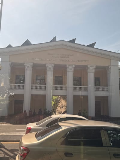 The image shows the front of a building with the inscription "O'ZBEKISTON RESPUBLIKASI ADLIYA VAZIRLIGI TOSHIKENT DAVLAT YURIDIK UNIVERSITETI" which translates to "REPUBLIC OF UZBEKISTAN MINISTRY OF JUSTICE TASHKENT STATE UNIVERSITY OF LAW".
The building has a classical architectural style with white columns and balustrades. The photo appears to have been taken from inside a car, as part of the vehicle is visible on the right side of the frame.
The location may be near the city of Kashgar mahalla, Uzbekistan, considering the sign's Uzbek language. However, the building itself is the Tashkent State University of Law in Tashkent, Uzbekistan.