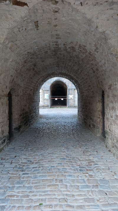 The image shows a view looking through a stone archway, likely part of a historic fortification or building in the city of Saint-Apollinaire, France. The tunnel-like passage is constructed from rough-hewn stone blocks. The floor of the passage is paved with cobblestones, which appear worn and weathered. On either side of the tunnel are narrow doorways, also made of stone. At the far end of the passage, another archway is visible, leading into a darker, more recessed area. A metal fence is positioned across this second archway, and some signs are mounted on the wall beyond it. The overall lighting suggests an outdoor environment, possibly during the daytime, with natural light filtering into the passage. There are no people or animals visible in the scene.