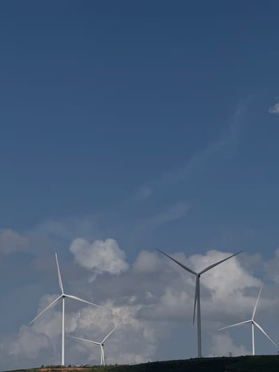 The image shows a landscape with wind turbines on a hill under a blue sky with scattered clouds. Four wind turbines are visible in the frame. The largest turbine is in the foreground on the left, with its blades slightly angled. To its right and further back, another wind turbine is visible with its blades also angled. In the distance, two more wind turbines are partially visible on the right side of the frame, with one closer than the other. The hill is covered with vegetation and appears to be a rural or semi-rural area. The sky is clear with some wispy clouds. The image appears to be taken from a low angle, looking up towards the wind turbines. The location is stated to be in the city of Da Tho, Vietnam.