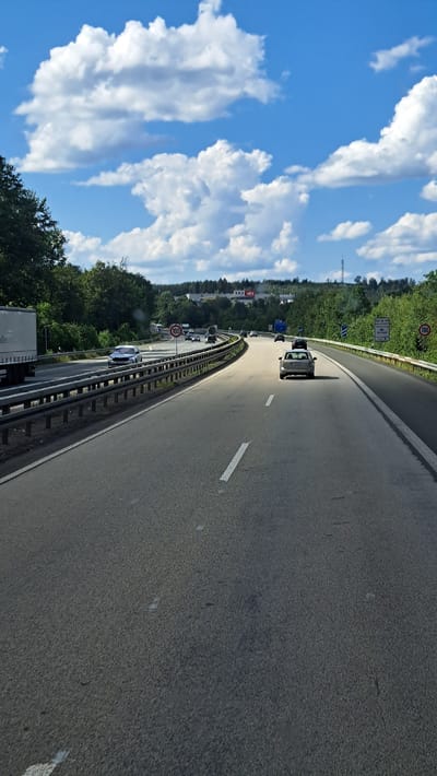 The image depicts a clear, sunny day on a multi-lane highway, likely in or near Freudenberg, Germany, given the presence of German speed limit signs. The perspective is from a vehicle traveling on the highway, looking forward.

On the left side of the road, a large white semi-trailer truck is parked or moving slowly in the leftmost lane. Beyond the truck, trees and vegetation line the roadside. A metal guardrail separates the lanes from the opposite direction of traffic.

The highway itself has at least two lanes visible in the direction of travel. Several cars are on the road, some in the distance and some closer. A silver sedan is in the right lane ahead, and a dark-colored car is in the left lane further up. A speed limit sign indicating "100" is visible on a pole on the right side of the highway. Another "100" speed limit sign is visible in the distance on the left.

In the background, a dense line of trees forms a natural border. Above the trees, a hazy industrial area with buildings and some smokestacks is visible. A tall radio tower can be seen in the distance. The sky is a vibrant blue with scattered, fluffy white clouds. The overall impression is of a typical highway scene on a pleasant day.