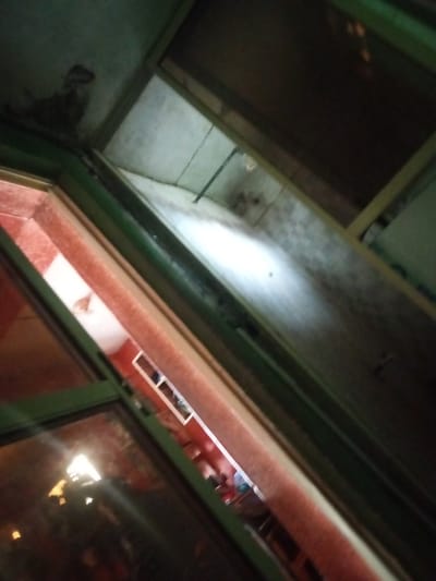 The image shows an interior view, looking upwards and diagonally into a space with architectural elements that suggest a multi-level structure. The dominant colors are dark greens, browns, and muted reds, with areas of bright light reflecting off surfaces.

In the foreground, there is a dark green framed window or opening. Through this opening, another layer of the structure is visible. This second layer appears to be a wall or partition with a reddish-orange textured surface. A bright light source is reflected in the glass or surface of this area, creating a strong highlight.

Further into the image, the perspective shifts upwards and into a darker, more recessed area. The walls here are a light, possibly white or grey, and show signs of wear or discoloration, with some dark patches suggesting mold or water damage. There are structural elements, possibly beams or framing, visible in the upper portion of the image, which are a light wood color.

In the mid-ground, within one of the openings, a small white object or structure is visible, possibly a vent or a recessed shelf. There are also reflections in the darker areas, indicating other light sources or objects present in the environment.

The overall impression is of an interior space that is partially lit and appears to be under construction, undergoing renovation, or in a state of disrepair. The angle of the shot and the lighting create a sense of depth and mystery.

Specific details about Agbara, Nigeria are not discernible from the image itself. The image is a visual representation of an interior architectural space.