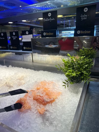 The image depicts a fish display at a market or restaurant in Herford, Germany. In the foreground, tongs are holding pieces of raw salmon on a bed of crushed ice. To the left, a white fish is also on display in ice. Behind the ice display, several black signs are visible with handwritten labels. One sign reads "Butterfisch," another "Lachs-filet," and another has the word "Zonder" on a white piece of paper. The signs also feature the Warsteiner logo. In the background, a person wearing a red top is visible, appearing to select fish. The environment appears to be an indoor setting with bright, artificial lighting, including blue LED lights overhead.
