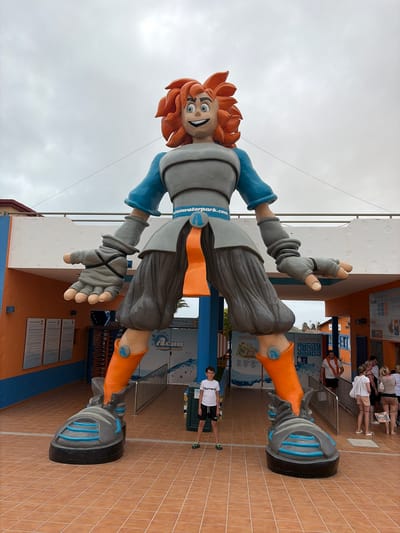 A large, colorful statue of a cartoon character stands prominently in the center of the frame, acting as a mascot for a water park. The character has spiky orange hair, wide blue eyes, and a big, welcoming smile. It wears a grey top with blue long sleeves, grey pants, and large orange and grey boots that resemble futuristic roller skates or hoverboard shoes, each featuring a blue "A" logo. A blue belt with "aquawaterpark.com" written on it cinches its waist. The character's hands are depicted with grey gloves.

In the background, several people are visible, some walking and others seemingly queuing near entrance gates or information booths. One young boy wearing a white t-shirt with "Money" printed on it and black shorts stands near the base of the statue on the right. The ground is covered in reddish-brown square tiles. Behind the people and statue are various buildings and structures, some blue and white, with signs and information boards mounted on the walls. One sign clearly shows "Aqua" and "Aquawaterpark" text, reinforcing the location. Another sign mentions "Priority Wristbands."

The sky above is overcast with light grey clouds, indicating daytime. The scene appears to be an outdoor public area, likely the entrance or a plaza of a water park, given the statue and signage.