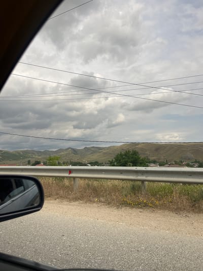 The image captures a view from inside a vehicle, looking out towards a landscape. The foreground shows a portion of the car's interior, including the side mirror and the asphalt road. A silver guardrail runs horizontally across the middle of the frame, separating the road from a dry, grassy embankment dotted with small yellow wildflowers.
Beyond the guardrail, a village or town is visible nestled at the foot of rolling, dry, grassy hills. The houses have reddish-brown roofs. The hills are undulating and appear arid, with sparse vegetation. In the background, the sky is overcast with dense, grey clouds. Several dark power lines are strung across the upper portion of the image, originating from the left side and extending towards the right. The overall scene suggests a rural or semi-rural setting under a cloudy sky, likely in the region of Karabudakhkent, Russia, given the context. There are no people or animals clearly depicted in the image.