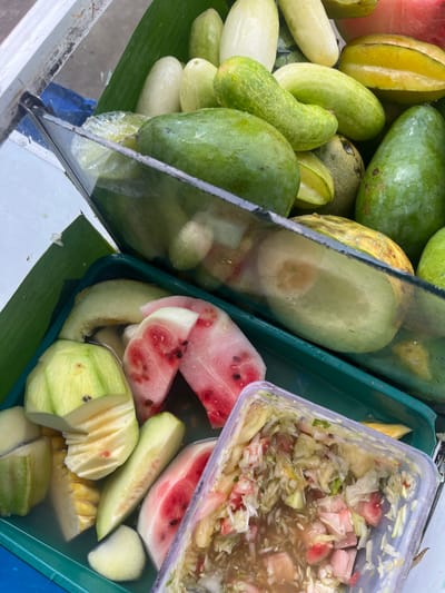 This is a close-up, high-angle shot of a fruit stand in Meteseh, Indonesia. The fruit stand is organized with various fruits displayed in green plastic trays and a clear plastic container. In the upper section, several green mangoes, cucumbers, and starfruits are visible, some covered with plastic wrap. The lower section contains sliced watermelon, pineapple, and other unidentifiable fruits in a green tray filled with water. A clear plastic container holds a mixture of chopped fruits, likely a fruit salad. The background is out of focus but suggests an outdoor setting, possibly a street vendor. The overall impression is of a fresh and abundant fruit selection offered at a local market.