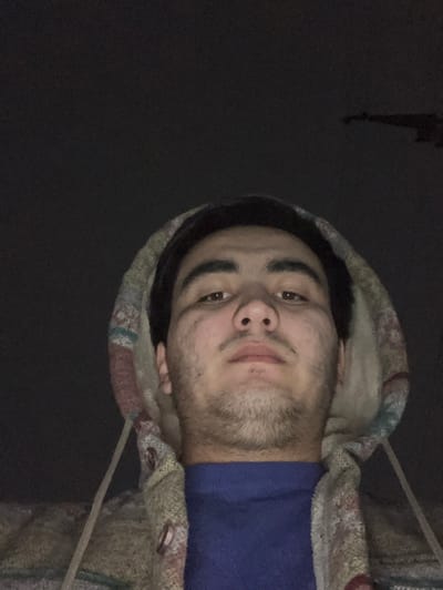A single male subject is captured in a low-angle, upward-facing photographic portrait, consistent with a selfie, against a dark backdrop. The subject is a young adult with dark hair, dark eyes, and light facial stubble, presenting a neutral expression while looking directly into the camera. He is dressed in a blue crew neck shirt beneath a hooded jacket featuring a multi-colored patterned fabric, including hues of beige, brown, red, and blue. The jacket's hood is up, framing his head, and visible drawstrings and buttons are present on the garment's front. The scene is dimly lit, with the subject's face illuminated by an apparent light source, possibly a camera flash or screen. The background is predominantly dark, revealing only a faint, indistinct, elongated dark structure in the upper right quadrant, potentially an architectural element or antenna, against what appears to be a night sky. No definitive landmarks are present within the frame to specify a precise location within Namangan City, Uzbekistan.