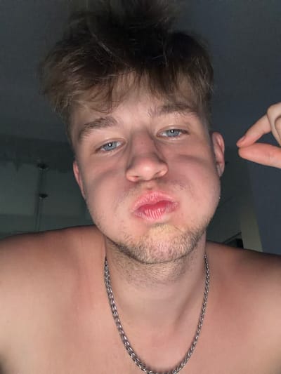 This is a close-up, selfie-style portrait of a young man with light brown, tousled hair and blue eyes. He is shirtless, wearing a silver chain necklace. His cheeks are puffed out, and his lips are pursed, creating a playful or exaggerated expression. His right index finger is visible near his right cheek, possibly indicating he is the one taking the photo. The background appears to be an indoor setting with neutral-colored walls, suggesting a home environment. There is no indication of the city of Vanino, Russia, in the image itself; this information is external to the visual content.