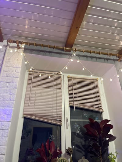 The image depicts an interior scene, likely a room, with a focus on a window and its decorations. The window is divided into two sections, each covered by light-colored horizontal blinds. Above the window, a string of small, warm-toned fairy lights is draped across a wooden curtain rod, extending from left to right. The ceiling appears to be paneled with light-colored planks, supported by a wooden beam. To the left of the window, a white brick wall is visible, illuminated with a purplish light. Inside the window, on the right side, a plant with dark red leaves is positioned. The overall impression is one of a cozy, decorated indoor space. The location is stated to be in the city of Mineralnye Vody, Russia. There are no people visible in the image.