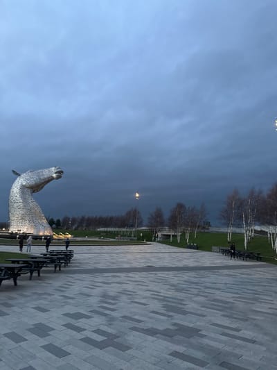 The image displays an outdoor plaza at The Helix, Falkirk, United Kingdom, under an overcast, dark grey sky, indicating dusk or early morning. The central subject on the left is one of "The Kelpies," a monumental sculpture of a horse's head constructed from interlocking metallic plates, illuminated from its base. The foreground consists of a patterned paved surface with various shades of grey rectangular tiles. Multiple dark-colored picnic tables with integrated benches are arranged on this paving and on a grassy bank to the right. Approximately five individuals are observed standing near the base of the sculpture on the paved area. A single person in dark clothing stands near a group of picnic tables on the right side, adjacent to a grassy slope. In the mid-ground, a tall street light with a bright yellow-orange luminaire is active. The background features a landscape of bare-branched trees, some with light-colored trunks, bordering a grassy area and obscuring a distant concrete structure resembling a bridge or underpass.