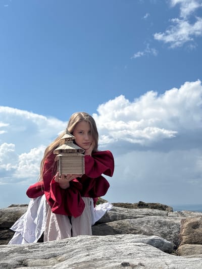 A young Caucasian female with long blonde hair is sitting on a rocky outcrop, holding a miniature wooden house. She is wearing a red top with full sleeves and a white skirt. Her left hand is supporting the wooden house, while her right hand is resting on her cheek. The background consists of a blue sky with white clouds and a distant landscape. The scene appears to be set in a natural, outdoor environment, possibly on a mountain or high plateau. The location is identified as Gubakha, Russia.