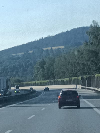 This media file depicts a view from a car driving on a highway in Hvězdonice, Czechia. The scene focuses on the road ahead, with several vehicles visible. In the foreground, a dark-colored SUV is driving away from the camera, its brake lights illuminated. Further up the highway, other cars are present, including a truck on the left side of the frame.
The highway has multiple lanes, indicated by white lines marking the boundaries. A barrier separates the lanes from the opposite direction of traffic. Beyond the highway, a lush green hillside is visible, covered in trees. Patches of yellow are scattered across the hillside, possibly indicating agricultural fields or flowers. The sky is clear and a light blue, suggesting it is daytime, likely late afternoon or early evening given the soft light. The overall impression is of a typical drive on a scenic highway in a rural or semi-rural setting. No discernible text is visible in the image.