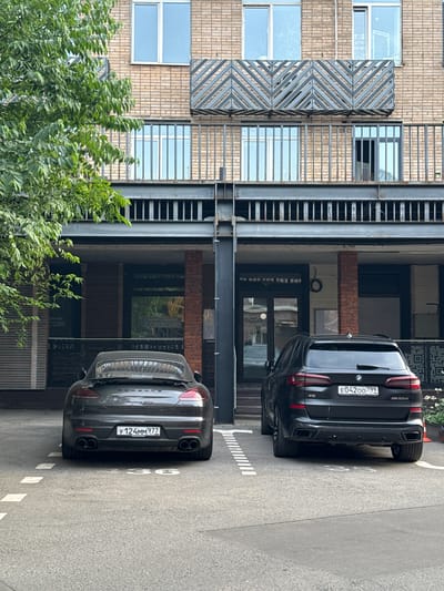A dark grey Porsche Panamera GTS and a black BMW X5 M50d are parked side-by-side in a parking lot in front of a modern brick building. The Porsche is on the left, with its rear facing the camera. The BMW is on the right, also facing away from the camera. The building has multiple floors with balconies featuring metal railings and decorative patterns. The ground floor has large windows and entrances, with some signage visible, including "caracole" and "cole". The parking lot surface is asphalt with white painted lines marking the parking spaces. A large green tree with dense foliage is on the left side of the image, partially obscuring the building. The license plate of the Porsche reads "Y124MM977" and the BMW reads "E04200799". The scene appears to be a street-level view in a city.