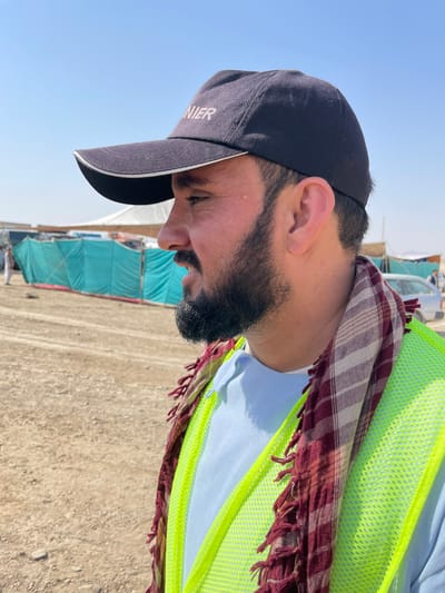 The image depicts an adult male in Pir Dust, Afghanistan, viewed in profile, facing right. He wears a dark blue baseball cap with "NIER" visible on the front, a light blue polo shirt, and a neon yellow mesh safety vest. A red and white checkered keffiyeh with fringe is draped around his neck. He has dark hair, a dark beard, and a short goatee. A small blemish is present on his right cheekbone. His mouth is slightly open, suggesting speech or an imminent vocalization. The background consists of dry, light brown unpaved ground. Behind him, a teal/green tarp structure forms a barrier, with white tents or shelters and faint human figures visible further in the distance. A light-colored vehicle is partially discernible in the far right background. The sky is clear and light blue, indicating bright daylight conditions.