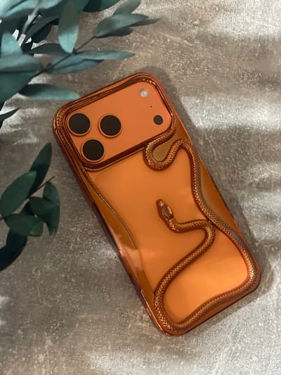 A close-up, high-angle shot shows an orange iPhone case with a metallic snake design on a textured grey surface. The case is positioned diagonally, with the camera lenses at the top left and the bottom right corner extending out of frame. The snake wraps around the case, its metallic texture catching the light. A sprig of dark green foliage is visible in the top left corner of the frame. The surface appears to be a rough, concrete-like material, with some lighter speckles.