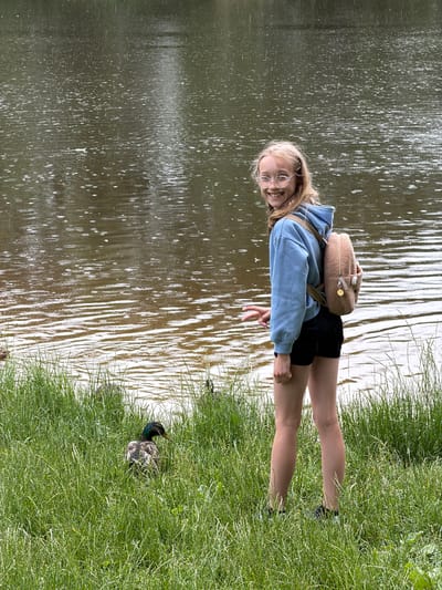 A young girl with blonde hair and glasses smiles at the camera, wearing a light blue hooded sweatshirt, black shorts, and a light brown fuzzy backpack. She is standing on the grass next to a body of water, possibly a lake or pond. The water has ripples and some reflections. A duck with dark and patterned feathers is resting in the grass near the water's edge. The scene appears to be set in a park or natural area, with lush green grass and the calm water suggesting a peaceful outdoor environment. The lighting suggests it is daytime, and the weather seems pleasant. The overall mood is relaxed and happy, with the girl's smile conveying a sense of enjoyment. The location is identified as Ulbroka, Latvia.