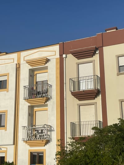 The image depicts the upper floors of an apartment building in Gafanha da Nazaré, Portugal, under a clear blue sky. The building's facade features a combination of cream-colored walls with decorative yellow and brown trim. There are multiple windows, some with white roller shutters. Two balconies are visible: one on the left, with a white railing and yellow decorative base, and another on the right, with a black railing and brown decorative base. The balconies appear empty or sparsely furnished. Lush green foliage from trees partially obscures the lower portion of the building on the right. The architectural style suggests a modern residential building.