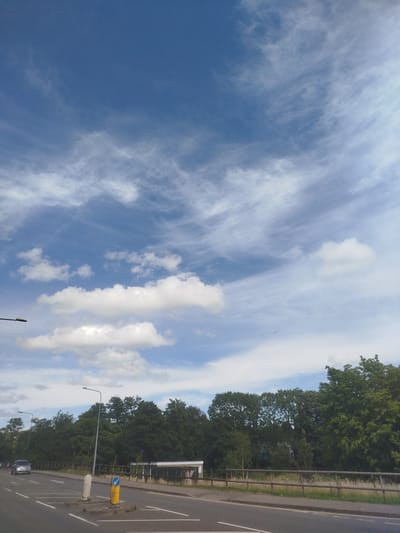 A wide-angle, eye-level shot depicts a road leading into a treeline under a vast blue sky filled with wispy white clouds. The sky occupies the majority of the frame, with the clouds appearing to stretch across the expanse, some denser and whiter, others thinner and more translucent, creating a dynamic and textured effect.
In the lower portion of the image, a two-lane road stretches from the left foreground towards the background. A silver car is visible driving on the left lane. Along the right side of the road, a wooden fence borders a grassy area. Behind the fence, a dense line of various green trees forms a natural barrier, with some interspersed buildings or structures visible through the foliage. Two streetlights stand on poles near the road, one in the mid-ground and another further back. A yellow and blue road sign is also present on the right side of the road.
The scene suggests it is daytime, likely late morning or afternoon, given the bright sky and clear visibility. The presence of clouds indicates a potentially changeable weather pattern, though the overall impression is of a bright day. The landscape appears to be a suburban or semi-rural area in Cwmbrân, United Kingdom, characterized by the road, streetlights, and abundant greenery. There is no discernible text visible in the image.