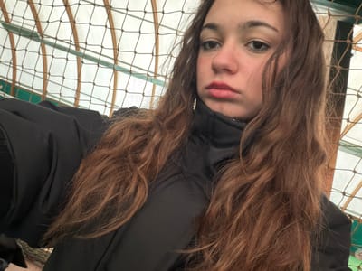 A young female individual is depicted in a close-up, angled portrait, likely a selfie, within a structure in Souffelweyersheim, France. She has long, wavy, reddish-brown hair, parted on the left, cascading over her shoulders and chest. She is wearing a black, high-collared puffer jacket. Her face shows dark eyes, defined eyebrows, and lips with red lipstick set in a slight pout, directed towards the camera. A dark mesh net is visible in the immediate background, with curved, light-brown architectural elements, possibly wooden beams, forming an arc pattern against a light-colored ceiling or wall. Horizontal green structural components are also present in the background. The environment appears to be a covered recreational or public facility.