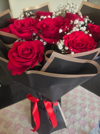 A bouquet of red roses and baby's breath is presented in a black and brown paper wrapping, tied with a red ribbon. The bouquet is placed on a table with a patterned cloth, possibly in Deira, United Arab Emirates. The lighting suggests an indoor setting.