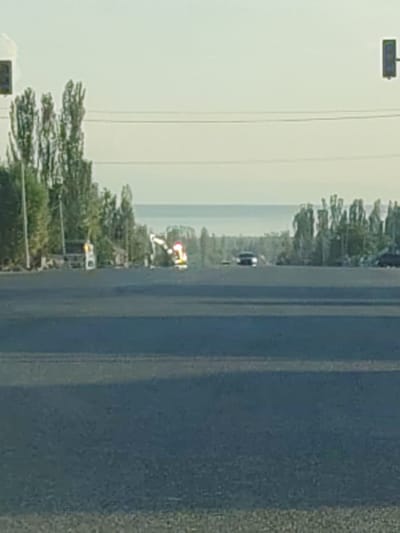 The image captures a street scene in Kachat, Kyrgyzstan, likely during the daytime with clear skies. The foreground is dominated by a dark asphalt road, suggesting a paved surface. In the middle ground, several objects and a natural landscape are visible. On the left, a white structure with yellow lights, possibly a small building or a roadside service area, is present, with a traffic light pole nearby. Further to the right, a vehicle with flashing red and yellow lights is observed, indicating a potential emergency vehicle or a hazard. Beyond this, a line of trees, predominantly tall and slender, forms a backdrop. In the distance, a body of water, likely a lake, is visible, with a hazy horizon separating it from the sky. A few cars are seen on the road in the mid-distance. The overall impression is that of a quiet, possibly rural or semi-urban, road in Kyrgyzstan. No text is clearly discernible in the image.