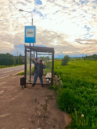 A woman in a blue jumpsuit and black vest stands inside a glass-enclosed bus stop on the side of a road near Курово, Russia. She is facing forward with her hands up against the glass, palms outward. To her left is a metal trash receptacle. A road with double yellow lines runs parallel to the bus stop, and beyond the road is a grassy area with trees. To the right of the bus stop, the ground is covered in green vegetation with dandelions. Above the bus stop is a blue sign with a white bus icon, mounted on a pole that also supports a street light. The sky is cloudy.