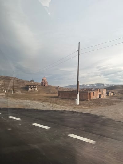 The image depicts a rural landscape in Voskepar, Armenia, viewed from a moving vehicle. The foreground features an asphalt road with white dashed lane markings, exhibiting horizontal motion blur. In the midground, dry, brown-hued terrain occupies the scene. To the left, a two-story stone building with a dark roof stands on a gentle slope, with multiple utility poles extending further up the incline. Dominating the center-right midground is a tall utility pole with several overhead wires. To the right, a cluster of unfinished or dilapidated brick and stone structures with open window/doorways is visible.
In the background, a distinctive red-domed church with an octagonal drum and conical roof, identified as the Holy Mother of God Church, is prominently situated atop a hill. Beyond this, a series of rolling, dry hills stretches towards the horizon, where distant mountains with snow-capped peaks are discernable. The sky is overcast, characterized by varying shades of grey clouds. No individuals or active interactions are visible. Reflections are present in the upper portion of the image, indicative of a shot taken through glass.