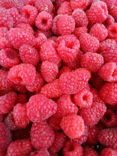 A close-up, overhead shot features a dense pile of ripe raspberries. The berries are a vibrant pinkish-red, with visible drupelets and a slightly fuzzy texture. Some berries are whole, while others appear to be broken or compressed from the weight of the pile. The lighting is bright, highlighting the natural sheen of the fruit. There is no discernible background or environmental context visible, suggesting the photograph was taken specifically to showcase the raspberries, likely in a food-related setting or as part of a larger collection of produce. The location in Кош-Дөбө, Kyrgyzstan is not visually represented in the image.