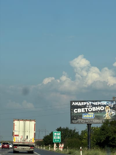 The image shows a highway scene near Yana, Bulgaria. In the foreground, a white truck with a red stripe drives on the right lane of the road. The truck has multiple speed limit stickers on its rear, indicating 80, 90, and 40. To the right of the road, a green highway sign indicates a parking area for trucks and a distance of 5 km. Further down the road, a large billboard is visible, advertising "ЛИДЕРЪТ ЧЕРПИ И НА СВЕТОВНО" with the "inbet" logo and an image of a soccer trophy and ball. The sky is partly cloudy. The environment appears to be rural or semi-rural, with trees and vegetation visible in the background.