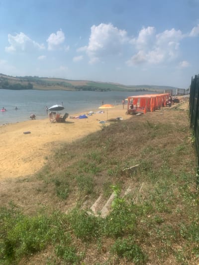 The image captures a sunny day at a beach along a river. The scene is populated with several people enjoying the water and the sand. Some are swimming, while others are relaxing on the beach, with a few under umbrellas. An orange tent-like structure, possibly a beach bar or seating area, is visible near the edge of the beach.
The weather appears to be warm and clear, with a bright blue sky dotted with white, fluffy clouds. The sun is shining, casting shadows and illuminating the sand and water. The river is calm and blue, with a grassy bank on the opposite side. The beach itself is sandy, with some areas of dry grass growing closer to the embankment. The embankment is covered in a mix of dry grass and green foliage, with a visible set of concrete stairs leading up from the beach.
The overall atmosphere seems relaxed and recreational, with people enjoying leisure activities on a summer day. The time of day is likely midday or early afternoon, given the bright sunlight and clear sky. There is no visible text in the image.