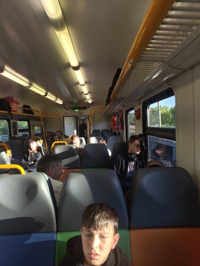 The interior of a passenger train car is shown, likely in Konobeevo, Russia, with several passengers seated and one standing. The car has a light-colored ceiling with fluorescent lighting, yellow handrails along the top, and rows of dark gray and blue-and-orange upholstered seats. Sunlight streams in from windows on the right side, casting shadows.
In the foreground, a young boy with brown hair and a dark hoodie is looking directly at the camera. Behind him, a man with gray hair and a white shirt is seen in profile. To the right, a young man with brown hair, wearing headphones around his neck and a black jacket over a grey shirt, is leaning his head against the window, appearing to sleep. His reflection is visible in the window, which looks out onto trees and a blue sky.
Further back, several other passengers are visible. On the left, a woman with dark hair is wearing a denim jacket and looking to her left. A man with glasses is seated next to her. Another passenger with short, gray hair is visible in the background. Near the door at the back of the car, a woman in a dark top is standing. Red fire extinguishers are mounted on the wall near the windows. Red bags with Russian text are hanging from the luggage rack above some seats. A digital display above the doorway shows "06" and the word "ВЫХОД" (Exit). The overall scene depicts a typical journey on a Russian train.