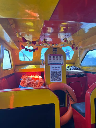 The image shows the interior of a small boat, likely a ferry or transport vessel, in the city of Tidore Islands, Indonesia. The boat's interior is painted in bright yellow and red. Along the ceiling, strings of small stuffed teddy bears and heart-shaped ornaments are hung. A poster with instructions on how to use a life jacket is displayed on a yellow panel, along with a QR code and the text "ARWANA MLU 10 NO. 540". A stack of orange life vests is visible on the left side of the boat. An orange life preserver ring with the word "ARWANA" printed on it is positioned in the foreground. Through a window at the back, a man wearing a blue shirt and shorts can be seen standing on the deck of the boat, facing away from the camera. The overall environment suggests a mode of transportation likely used for inter-island travel in the region.