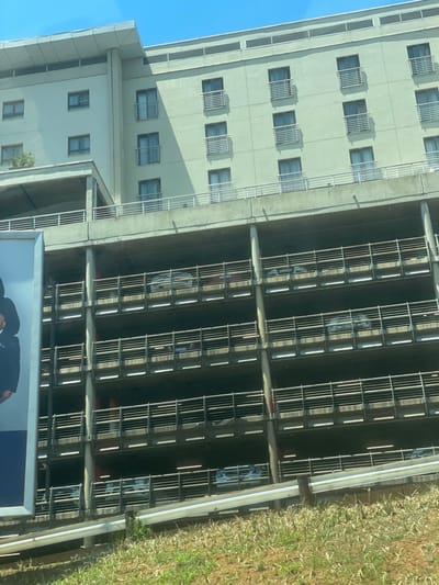 The image depicts a multi-story building complex in Kempton Park, South Africa, consistent with the architecture of the City Lodge Hotel Group properties near O.R. Tambo International Airport.
The scene features a modern, pale beige or light grey multi-story building with a flat roof, comprising at least seven visible floors. Each floor contains multiple rectangular windows, many equipped with small, grey horizontal-barred balconies. Shadows are cast on portions of the building, indicating bright daylight.
Directly beneath the main building is a concrete multi-level parking garage, at least five levels of which are visible. Each level of the garage is secured by horizontal metal railings. Numerous passenger vehicles, including light and dark-colored sedans and SUVs, are parked within the structure on various levels. Vertical concrete pillars support the parking garage.
On the far left, a large billboard or sign is partially visible, displaying the torso of an individual, likely male, wearing dark formal attire. The foreground of the image shows a grassy embankment with dry, light-colored vegetation. A silver metal guardrail runs horizontally across the lower section of the frame, positioned between the grassy area and the parking structure. The sky above is clear and blue. No individuals are visible performing actions or interactions within the scene.