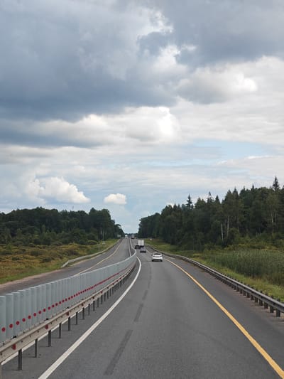 This is a vertical, eye-level, wide shot of a highway on a cloudy day. The road is asphalt with a yellow line marking the center and a white line marking the right lane. On the left, a guardrail with red reflectors runs along the side of the highway, separating it from a vegetated embankment. On the right, a metal guardrail lines the edge of the road, with dense green forest beyond it. The sky is filled with dramatic, dark gray clouds with patches of white and some lighter blue sky visible. Several vehicles, including cars and trucks, are driving on the highway in the distance. The scene appears to be in a rural or suburban area, possibly on the outskirts of Ямская Слобода, Russia, given the presence of a highway and forested landscape.