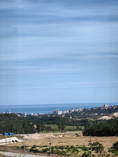 The image depicts a wide-angle view of a coastal landscape under a bright, partly cloudy sky. In the foreground, a dry, grassy field with some scattered bushes and barbed wire fencing suggests a rural or undeveloped area. Several utility poles stand in the field, one with a dark object attached. Further back, a road runs horizontally across the frame, with what appears to be construction materials, including piles of sand and what might be portable buildings, alongside it. There are also two concrete culverts visible.

Beyond the road, a lush, green hillside covered in trees slopes down towards the sea. Nestled on the hillside and along the coast is a densely populated city with numerous white, multi-story buildings, some appearing to be apartment complexes or hotels. The city stretches along the coastline, with a clear blue sea meeting the land. The sky is a pale blue with thin, horizontal clouds, indicating fair weather. The sunlight suggests it is daytime, likely late morning or early afternoon. There are no discernible people or specific activities taking place, giving the scene a sense of stillness. The location is identified as Kiparisnoe, Ukraine. The overall impression is one of a coastal city developing amidst a natural landscape.