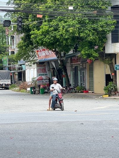 An individual wearing a white helmet, grey long-sleeved shirt, and blue jeans rides a red electric scooter down a street in Tân Quang Commune, Vietnam. The scooter has a black seat and front wheel. The individual's legs are visible, wearing light-colored sandals. The street appears to be paved with asphalt and has a yellow line dividing the lanes.

To the right of the scooter, a sign with "25B" is visible on a pole. In the background, several shops and buildings line the street. One shop has a large red and white sign with Vietnamese text. Another building has a white facade with green accents and large metal roll-up doors. Trees with lush green foliage overhang the street and buildings.

To the left of the scooter, a traffic light is visible, with three circular lights arranged vertically, and a black digital display to its left. A white truck with a grey tarp covering its load is parked on the left side of the street. Several motorcycles are parked in front of the shops. Two green trash bins are situated near a small red and white umbrella. The overall scene depicts a typical street view in a Vietnamese commune.