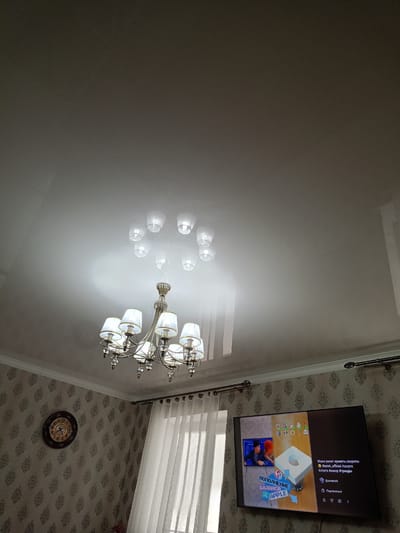 This media file captures an indoor scene, likely a living room, at night. The primary subjects are a modern chandelier hanging from a glossy, reflective ceiling, and a flat-screen television mounted on a wall. The television displays a video with Russian text, featuring graphics and text overlays that suggest content related to "top-ups" or "recharging" ("ПОПОЛНЕНИЕ"), "balance" ("БАЛАНСА"), and "Apple." There are also hashtags indicating humor and trends.
On the wall to the left of the television, a round analog clock with Roman numerals is visible, showing the time is approximately 7:05. This suggests the image was taken in the evening or early morning. The wall is covered in patterned wallpaper, and sheer white curtains are partially drawn over a window to the right of the clock. The overall lighting is dim, with the chandelier providing a warm, central light source and the television screen illuminating the immediate area. The reflective ceiling casts light and the chandelier's form in a diffused, ethereal manner, creating a somewhat atmospheric mood. The scene appears to be a domestic setting, possibly in Belidzhi, Russia, given the location context. No people are directly visible, but the content on the screen implies an ongoing engagement with digital media.