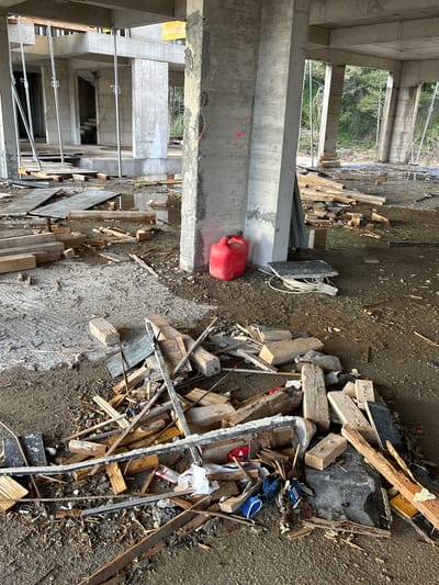 The scene depicts the interior of an unfinished concrete structure, likely a ground floor or basement, in Kavač, Montenegro. The environment is characterized by an unkempt construction site floor composed of dirt, scattered concrete fragments, and multiple puddles of standing water. Dominant features are robust, unpainted concrete pillars and beams forming the building's skeleton, extending into the background. Several metal adjustable shoring posts are visible in the mid-ground, supporting upper concrete slabs.

In the foreground, a substantial pile of discarded wooden planks, various timber scraps, and metal rebar lies on the dirt floor. Amidst this debris, smaller pieces of concrete rubble, plastic refuse, and what appears to be a blue fabric item are intermingled. To the right of the central foreground, a bright red, plastic jerry can rests upright at the base of a concrete pillar. Adjacent to the jerry can, a coil of white electrical cable and a small electrical junction box or device are placed on a dark, flat, rectangular surface, possibly a piece of roofing felt or similar material, near a puddle.

Beyond the concrete structure, a natural outdoor environment with green foliage and trees is visible, suggesting the building is partially open-sided or still under exterior development. No people or active construction actions are present in the image. The overall impression is one of a paused or abandoned construction phase, marked by disorder and accumulated debris.