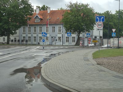 The image shows a street scene in Šilutė, Lithuania, on a cloudy day. The foreground features a wet, paved road with reflections of buildings and trees in puddles. A curved, grey paved sidewalk runs along the right side of the road. In the background, a light-colored building with an orange tiled roof stands, adorned with a Lithuanian flag. Several trees with green foliage are visible, some casting shadows on the building. Various traffic signs are present, including pedestrian crossings, lane direction indicators, a speed limit sign (30t), and a yield sign. Construction barriers and white tarpaulins are visible on the right side of the scene. The overall impression is one of an urban environment experiencing a recent rain.