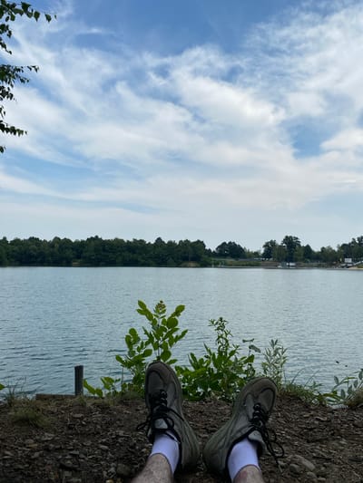 This media file is a photo that appears to be taken on a sunny day in Kaniów, Poland. The image is oriented sideways, with the horizon running vertically.
The scene depicts a body of water, likely a lake or river, with ripples on its surface. Along the edge of the water, there is a bank covered in dirt and small stones. Green vegetation, including leafy bushes and grass, grows along the bank. In the foreground, two feet wearing athletic shoes and white socks are visible, suggesting someone is sitting or standing on the bank, looking out at the water. The shoes are a muted grey color.
Across the water, a dense line of trees forms the far bank, with some structures and possibly a road visible in the distance amongst the trees. The sky above is a light blue, with wispy white clouds scattered across it, indicating fair weather. The overall impression is one of a peaceful, natural setting, possibly a park or recreational area, where someone is enjoying the view. There are no visible signs of text in the image.