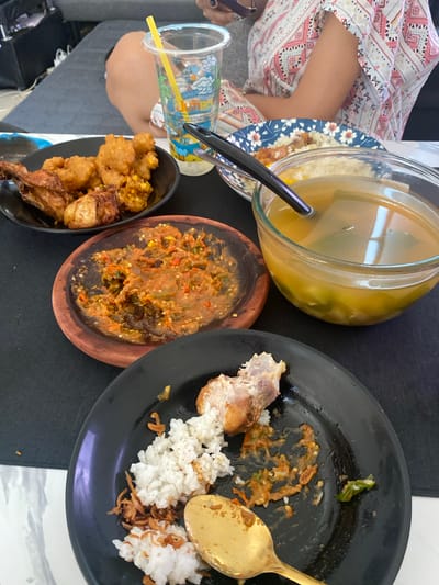 A overhead shot captures a table setting with a meal in progress. In the foreground, a black plate holds remnants of a meal, including white rice, a piece of fried chicken, and a dollop of red chili paste. A golden spoon rests on the plate. Adjacent to this, a small, rustic wooden dish contains more of the red chili paste, appearing to be a condiment. Further back, a clear glass bowl is filled with a yellowish broth, likely a soup, with some green vegetables visible within. Another plate, decorated with blue floral patterns, sits behind the soup, holding a piece of fried chicken and rice. To the left, a black bowl contains a serving of fried chicken pieces. A clear plastic cup with a colorful cartoon design and a yellow straw is also present, suggesting a beverage. The table is covered with a black tablecloth, with a portion of a white and patterned garment, likely worn by a person seated at the table, visible at the top. The background shows a blurred view of a gray sofa, indicating an indoor setting. The presence of Indonesian dishes like fried chicken and chili paste strongly suggests the location is in Indonesia, potentially in a casual dining setting in Cibinong, a city known for its culinary offerings.