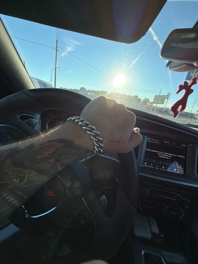 The image shows a first-person perspective from inside a car, likely in Pokrovskoe, Russia, on a sunny day. The driver's hand, adorned with a thick silver bracelet and a small red cross tattoo on the knuckles, is gripping the steering wheel. The steering wheel has a black leather finish with a red Dodge logo in the center. The car's dashboard features a digital display showing the time as 14:13 and the temperature as 20.0°C. Text on the screen, in Russian, appears to be related to phone connectivity. Outside the car, the bright sun is visible through the windshield, creating lens flare. Power lines and trees are visible in the distance, suggesting a semi-urban or suburban environment. A red, car air freshener shaped like an animal hangs from the rearview mirror. The sky is clear and blue.