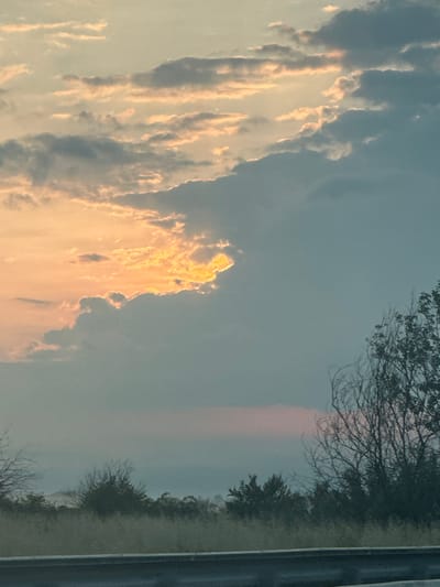 The image displays a vertical outdoor landscape, captured during what appears to be either sunrise or sunset. The left side of the frame is dominated by a vast expanse of sky filled with clouds. Many of these clouds are brilliantly illuminated with warm, golden, and orange hues, indicating the presence of the sun at a low angle. These brightly lit clouds contrast starkly with the cooler blue-grey tones of the less illuminated parts of the sky.
On the right side of the image, a darker segment of sky transitions downwards into a line of trees and dense foliage. These trees are largely silhouetted, with some bare branches discernible against the softer sky, suggesting late autumn or winter. Below the tree line, on the far right edge, a dark, straight, vertical strip is visible, likely representing a road or a guardrail alongside it. A faint, elongated reddish-pink glow is visible in the upper-right portion of the sky, above the tree line, possibly an atmospheric phenomenon like a sun pillar or a result of light scattering.
The overall impression is one of natural beauty and tranquility, with the dramatic lighting on the clouds being the focal point. There is no visible text in the image. The composition, with the sky on the left and terrestrial elements on the right, suggests the original photograph might have been taken with the camera rotated 90 degrees clockwise from a standard landscape orientation. The scene evokes a sense of calm and the natural splendor of twilight hours in a rural or semi-rural setting.