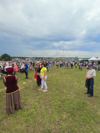 This image depicts an outdoor event, likely a festival or gathering, on a grassy field in the village of Koyanovo, Russia. A large crowd of people, consisting of men, women, and children, are present. Many are standing and observing, while some are seated on benches in the background. Several tents and stalls are visible in the middle ground, suggesting vendors or information booths. In the foreground, a person in traditional clothing stands with their back to the camera, facing the crowd. Another man in a light-colored shirt and jeans stands to the right, looking towards the viewer. The sky is overcast with dramatic clouds. The overall atmosphere appears to be that of a community event.