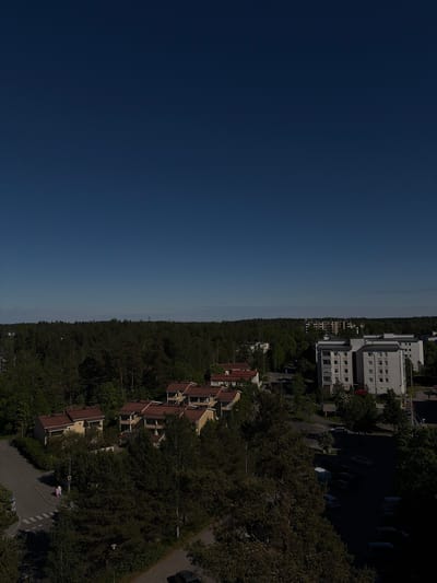 The image shows an aerial view of a suburban area, likely in Espoo, Finland. On the right side of the image are residential buildings, including apartment blocks and houses with reddish-brown roofs, interspersed with dense green trees. A parking lot with several cars is visible near the apartment blocks. A road winds through the residential area. On the left side of the image is a clear blue sky.