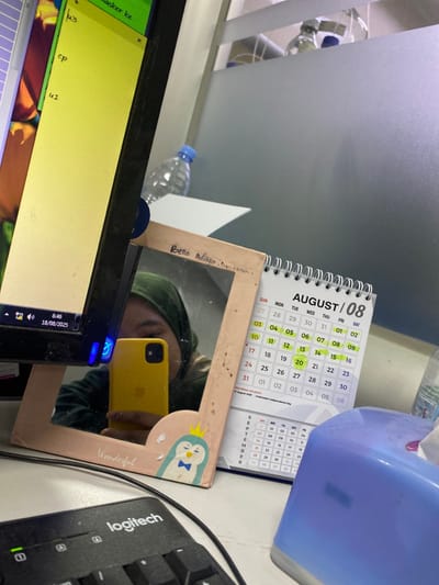 The scene depicts an interior office or cubicle environment, likely located in Indonesia, as indicated by a national holiday reference on a desk calendar. The view is from a slightly elevated perspective, focusing on a light-colored desk surface.
**Subjects:** A person is partially visible as a reflection in a small, wooden-framed mirror positioned on the desk. The reflection shows the lower portion of the person's face, covered by a dark green hijab, and a bright yellow smartphone held up, presumably to capture the image.
**Objects:** A black computer monitor occupies the left side of the desk, displaying a light yellow interface with text and a Windows taskbar that shows the time "6:46" and the date "18/08/2023." Adjacent to the monitor, the aforementioned wooden-framed mirror is visible. The mirror's frame is inscribed with "Bella Adilia Sultana" at the top and decorated with the word "Wonderful" and a cartoon penguin graphic at the bottom. To the right of the mirror, a white desktop calendar for "AUGUST / 08" (August) stands upright. Several dates on the calendar are highlighted in yellow, and text below the main calendar section notes "INDONESIA HOLIDAY: 17 August 2023 (Independence Day)." A black Logitech keyboard is partially visible in the bottom left foreground, and a blue tissue box is partially visible on the far right.
**Actions/Interactions:** The person is actively taking a photograph of their reflection using the yellow smartphone, with the computer monitor and calendar forming part of the background.
**Environment:** The background features a frosted glass partition or wall. Two clear plastic water bottles with blue caps are visible on a surface behind the desk, through the partition. The area is illuminated by artificial light sources.
**Location Detail:** While the calendar confirms the setting is in Indonesia, there are no visual cues within the image to specifically identify the city of Cipeundeuy.