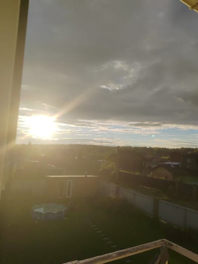 The image depicts a view from a window overlooking a rural or suburban area, likely in Zhal'skoe, Russia, at sunset. The sun, a bright, lens-flared orb, is positioned in the upper left quadrant of the sky, casting a strong glare and illuminating the clouds and landscape below. The sky is a mix of dark, overcast clouds and patches of lighter, more scattered clouds, with a visible sunbeam cutting through.

In the foreground, on the left, a portion of a white window frame or wall is visible, and a light-colored wooden railing is present at the bottom right. Below the window, a green lawn occupies the lower half of the image. On the lawn, a round, above-ground swimming pool with blue-grey sides is situated. Adjacent to the pool, a small, rectangular building with two large, light-colored doors or windows is visible.

The middle ground reveals a cluster of houses with dark-colored roofs and visible windows, nestled amongst trees. A light-colored fence runs along the edge of the property in the middle right. The background consists of a distant tree line against the horizon. The overall atmosphere is that of a tranquil evening setting, with the strong sunlight creating a dramatic effect. There are no people or animals clearly visible in the image.