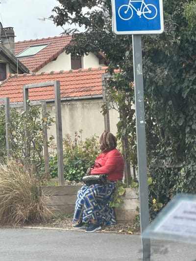 An individual is seated facing left, positioned next to a wooden raised planter or border on a paved surface in Stains, France. The person wears a red long-sleeved top, a long blue skirt with a yellow geometric pattern, black sneakers with white soles, and a multi-patterned head covering. A black handbag with a furry or textured trim is held in their lap. To the right, a grey pole supports a blue square sign featuring a white bicycle icon. Behind the seated individual is a garden area with various plants, including tall dry grasses, leafy green vegetation, and some reddish foliage, contained within the wooden structure. Further back, residential buildings are visible, characterized by red tiled roofs, a skylight, and a light-colored stucco wall. Overcast skies are present.