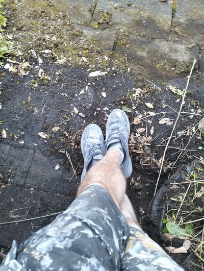 A person's legs, wearing grey camouflage-patterned shorts splattered with white and tan paint, and grey athletic shoes with grey laces, are positioned on a surface of dark, damp soil mixed with small stones, scattered leaves, and debris. The ground also appears to be partially paved with grey interlocking bricks, some of which are covered in a thin layer of dirt and moss. The scene is outdoors, with dry grass stalks and a few green plants visible. The context of the image suggests it was taken in Enschede, Netherlands, possibly in a garden, yard, or construction area, given the presence of paint splatters and the unkempt ground.